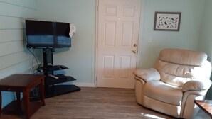 A living room with a flat-screen TV on a stand, a beige leather recliner, a wooden coffee table, and a white door. - Clean spacious 2BR mobile home in great senior community in Largo Florida (Largo)
