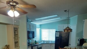A dining area with a wooden table and floral chairs, a ceiling fan, and a kitchen in the background. - Clean spacious 2BR mobile home in great senior community in Largo Florida (Largo)