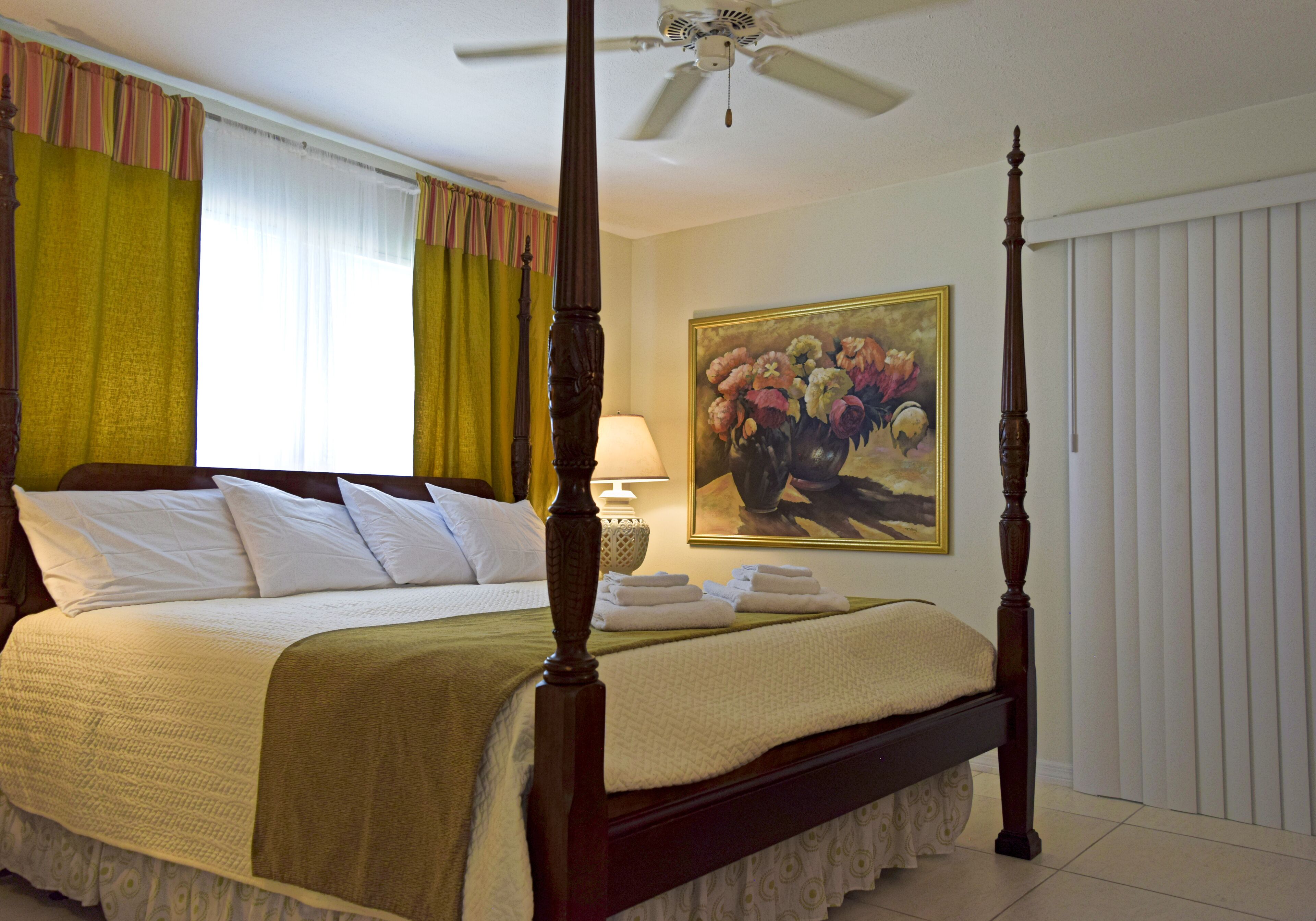A four-poster bed with a canopy, a painting of flowers on the wall, and a ceiling fan.