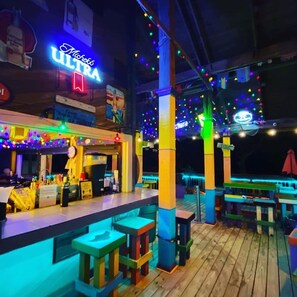 Ein Außenbereich mit Bar, beleuchtet durch farbiges Licht, mit einer Neonreklame und Holzsäulen.
