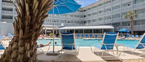Zona de piscina con sillas de descanso, una palmera y un edificio de varios pisos con balcones.
