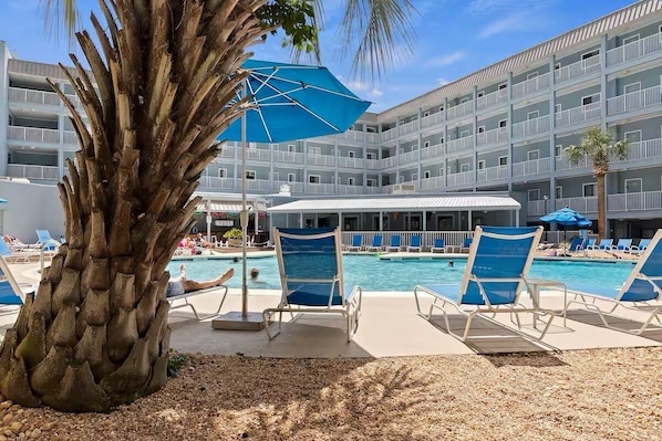 A pool area with lounge chairs, a palm tree, and a multi-story building with balconies.
