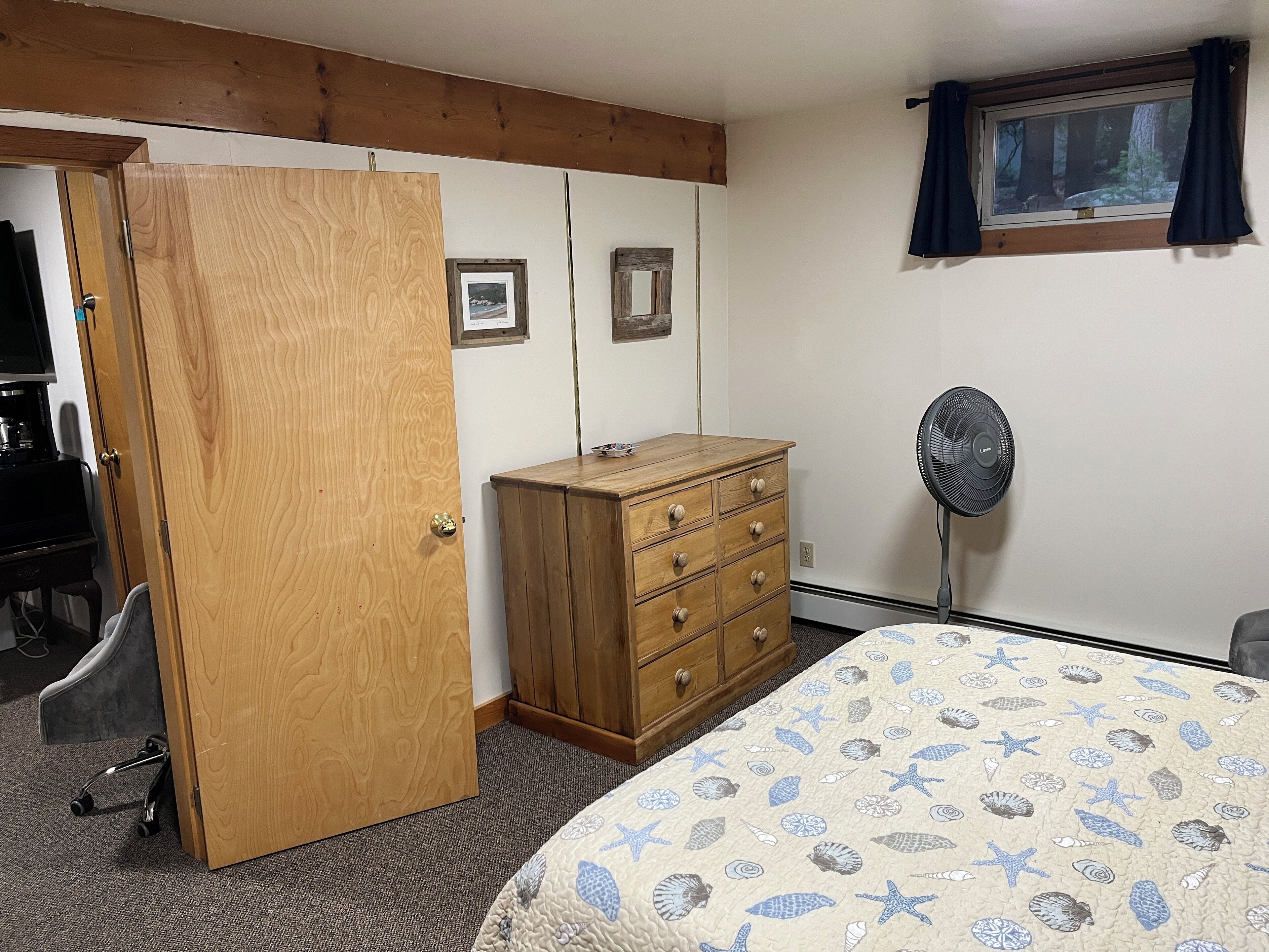 A bedroom with a wooden wardrobe, a bed with a patterned duvet, a standing fan, and a window with curtains.