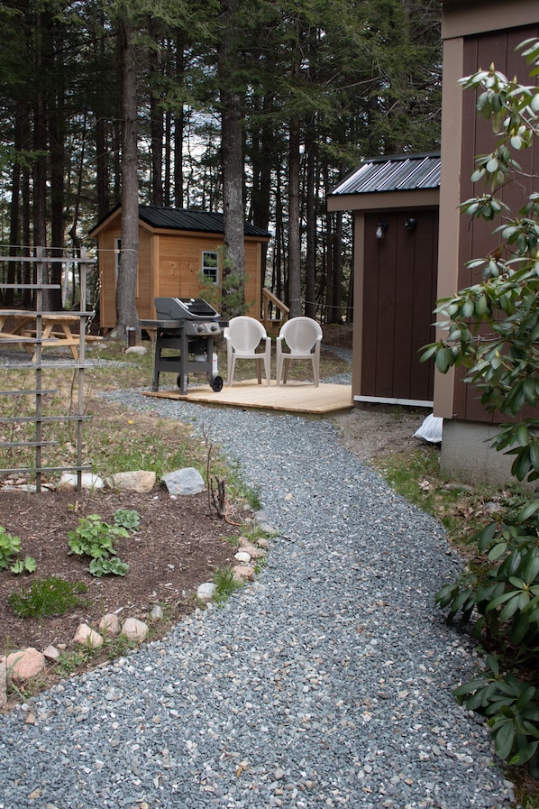 A gravel path leading to a wooden deck with two white chairs and a grill.