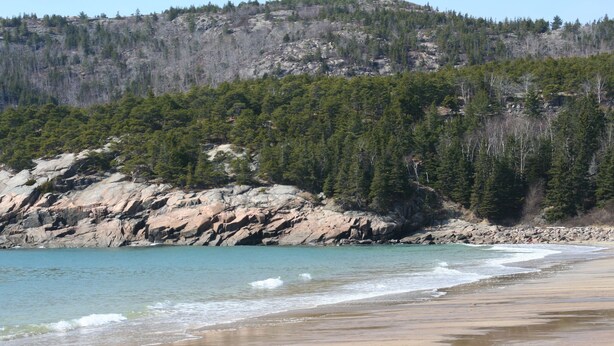 A beach with clear turquoise water, a sandy shore, and a forested hill in the background.