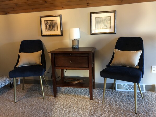 A room with two dark blue armchairs with gold cushions, a small wooden side table with a lamp, and two framed artworks on the wall.