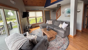 A spacious living area with a grey sofa, a wooden coffee table, and a patterned rug. A sign reading "BEACH HOUSE" is visible on the wall.