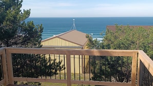 A wooden deck with a railing, overlooking a building with a sloped roof and a view of the ocean.