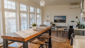 A spacious living area with a long wooden table, multiple seating options, and large windows providing natural light. - Haven on Ida - Lakeside Cabin, Cozy Porch + Sauna + Kayaks (Alexandria)