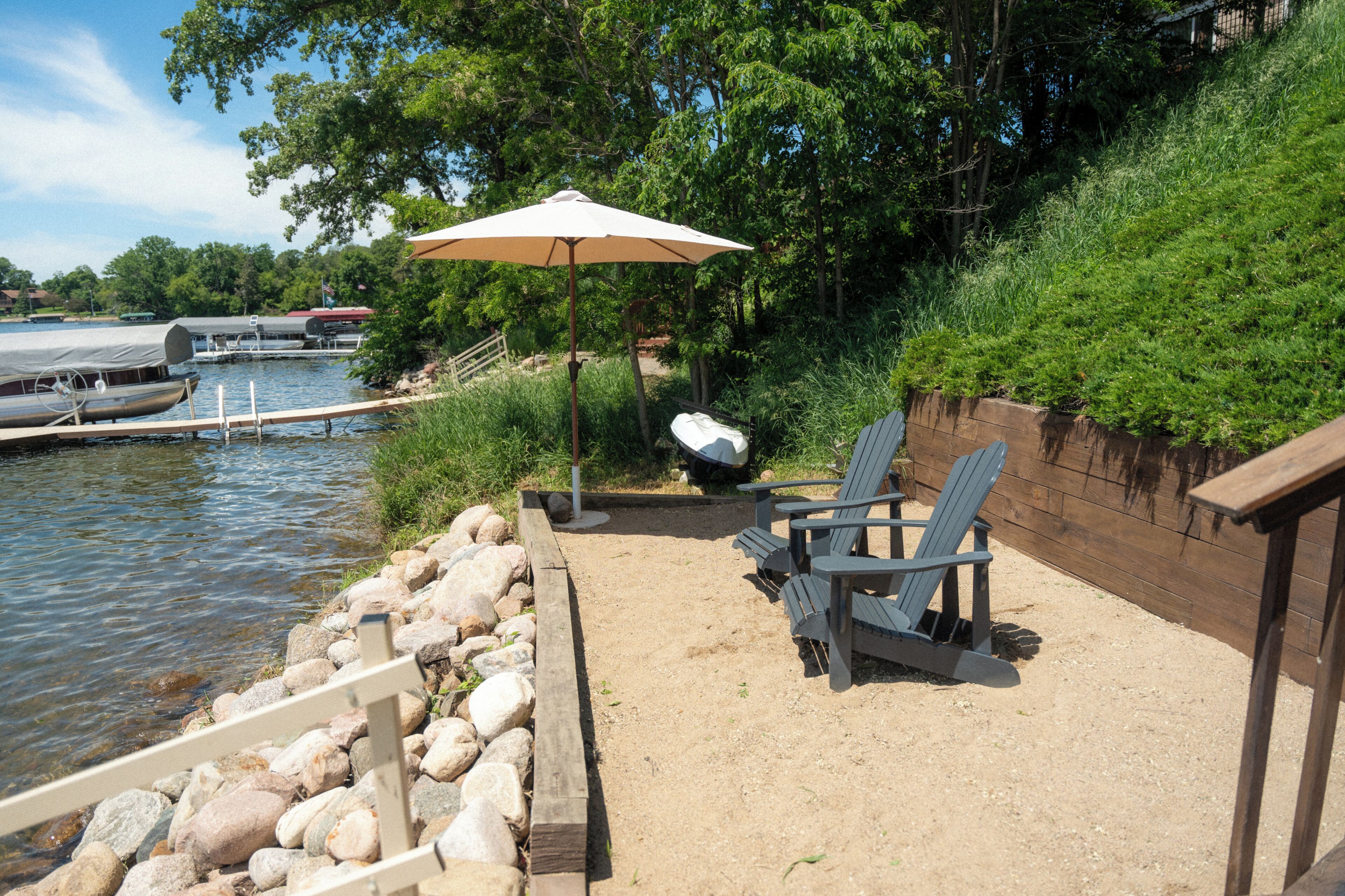 A lakeside patio with Adirondack chairs, a picnic table, and a large umbrella.