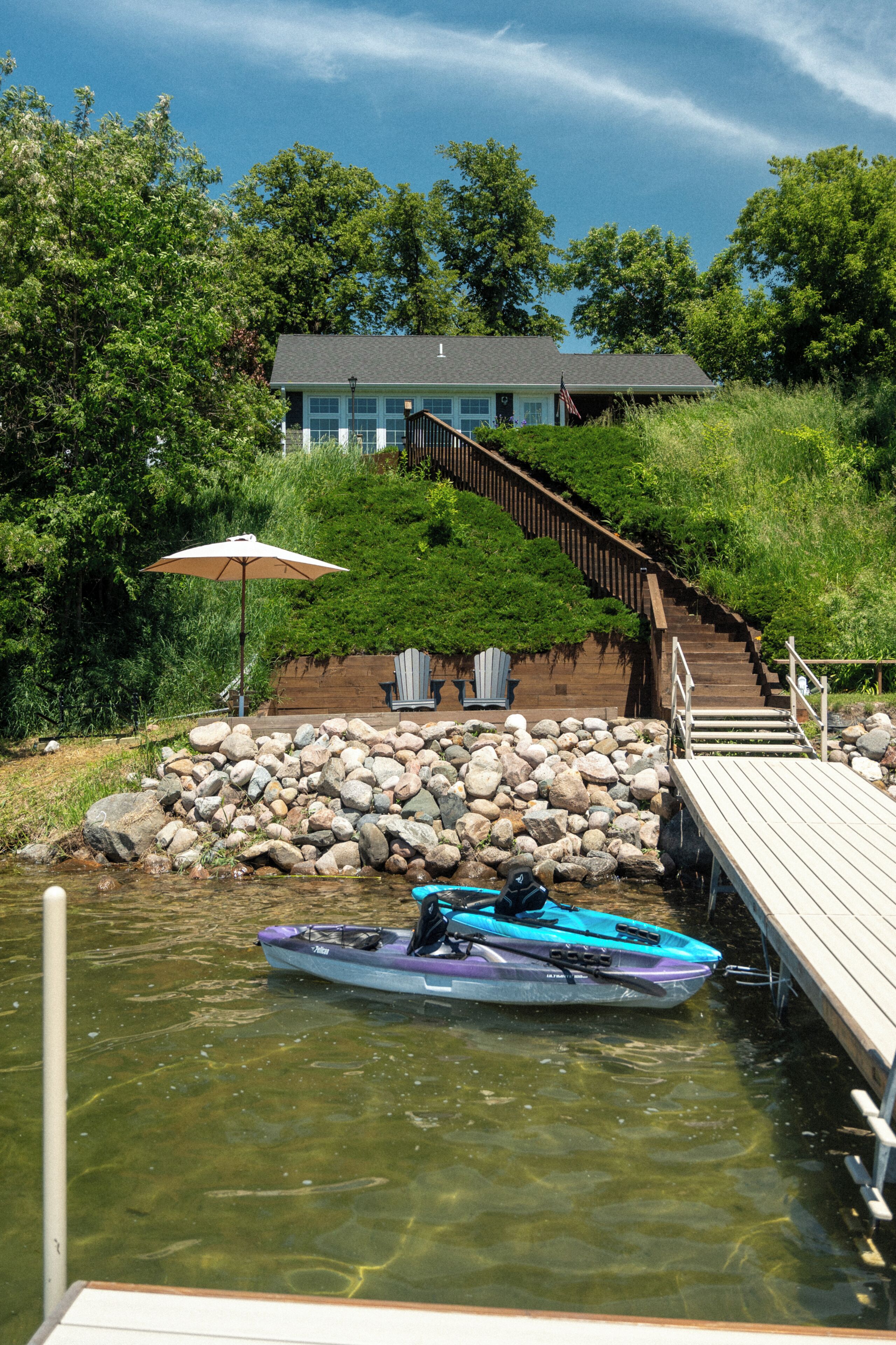A dock with a kayak, a wooden staircase leading to a house, and a stone wall with chairs.