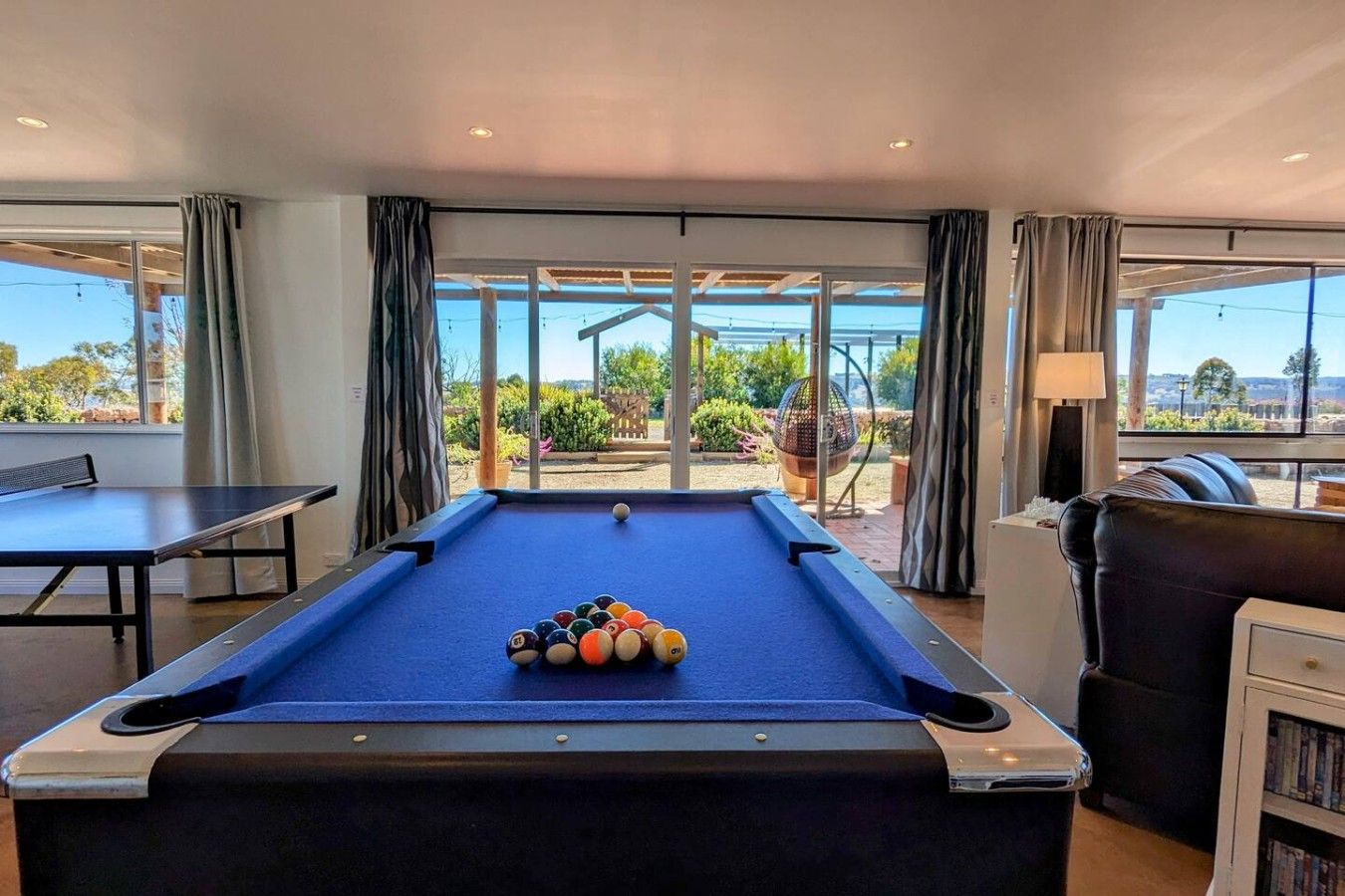 A pool table with balls in the center, a ping pong table to the left, and a sofa with a bookshelf to the right.