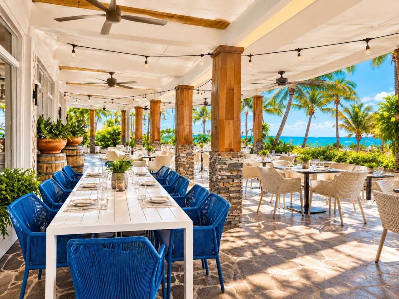 An outdoor dining area with a long table, blue chairs, and a view of the ocean.