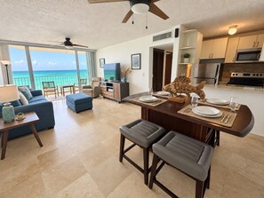 A modern living room with a large window overlooking a beach, a dining area with a wooden table and bar stools, and a kitchen with stainless steel appliances.