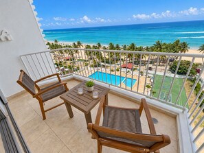A balcony with two wooden chairs and a small table, overlooking a beach with a pool and a tennis court.