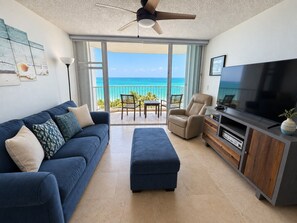 A living room with a sofa, ottoman, TV stand, and a balcony with a view of the ocean.