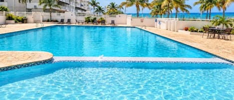 A clear swimming pool with a white deck, surrounded by palm trees and a building in the background.