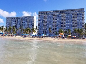 A beachfront hotel with multiple floors, palm trees, and beach umbrellas.