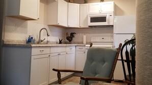 A compact kitchen with white cabinets, a microwave, a stove, and a sink. There's a wooden chair with a cushion and a small table with a plant. - Bungalo off the Withlacooche (Dunnellon)