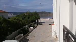 Ein schmaler Pflasterweg führt zu einer Uferzone mit Blick auf das Meer und Hügel in der Ferne.