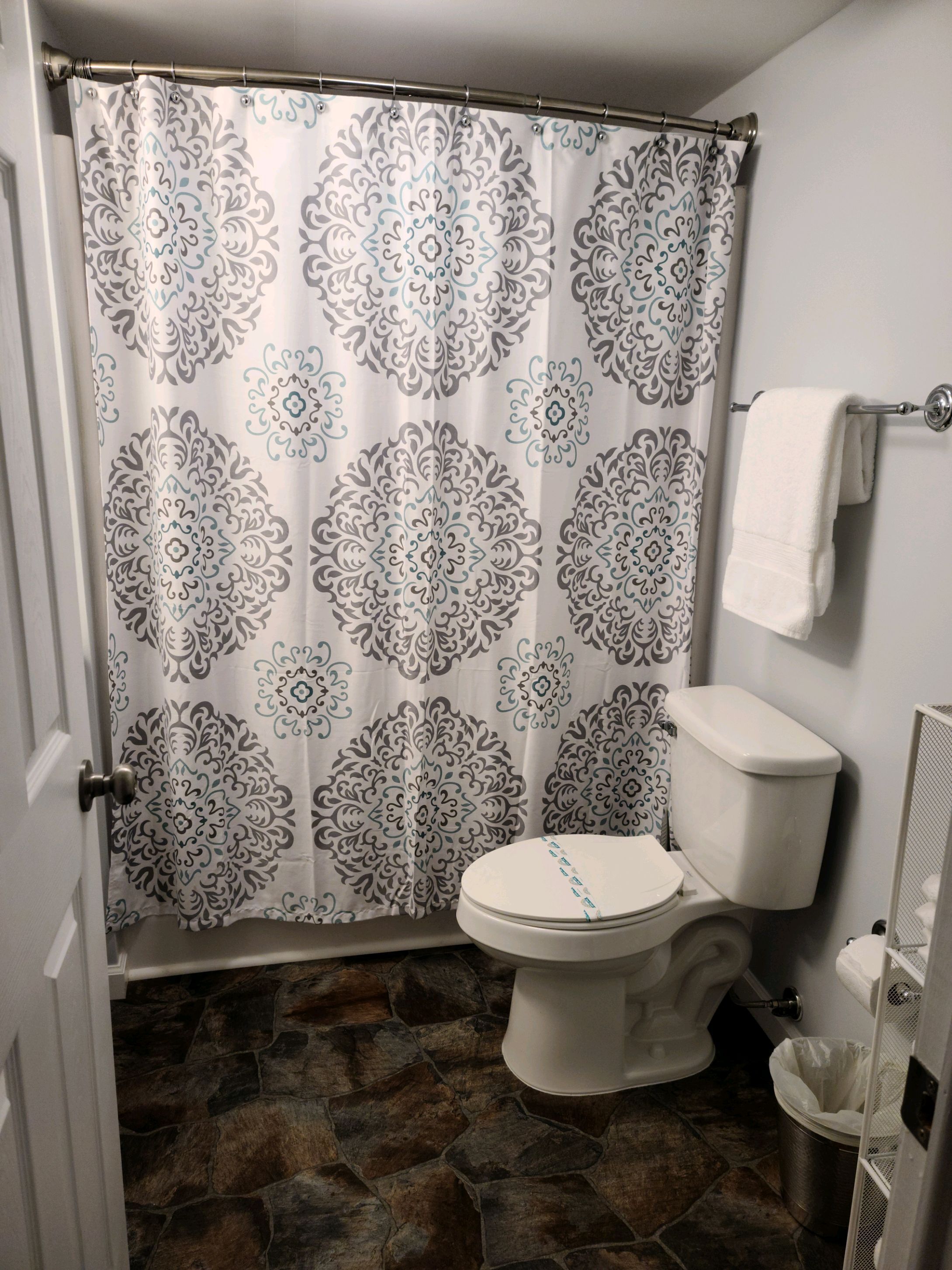 Un baño con cortina de ducha estampada, inodoro, toalla blanca en un perchero y suelo de baldosas de piedra.