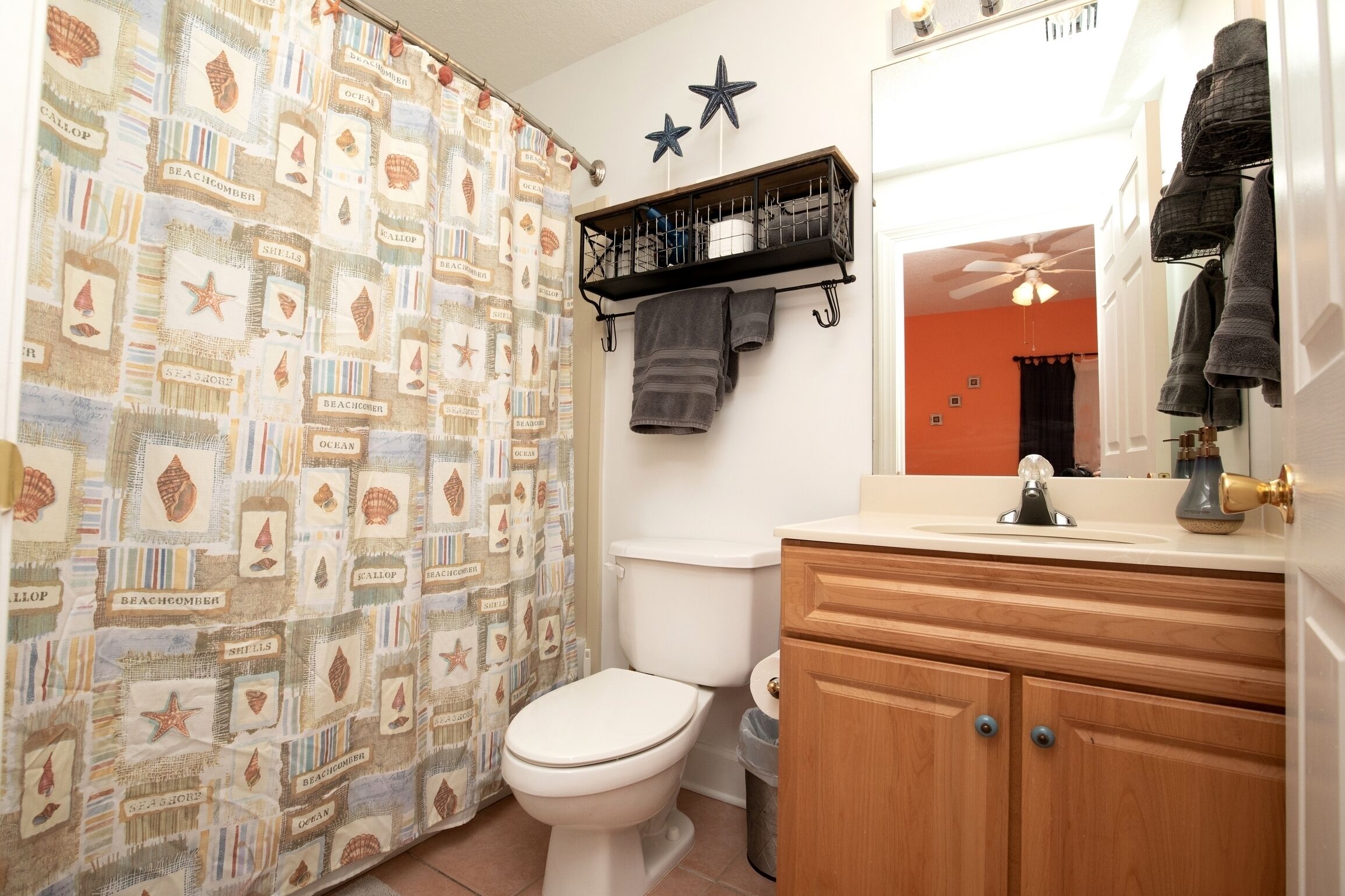 A bathroom with a nautical-themed shower curtain, a toilet, a wooden vanity with a mirror, and towels hanging on a rack.