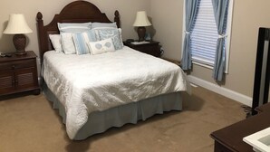 Una cama bien hecha con edredón blanco y detalles azules, flanqueada por dos mesitas de noche con lámparas, un aparador y una ventana con cortinas.