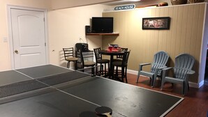 Una mesa de ping pong con dos paletas, un televisor y un cartel que dice "BEACH HOUSE" en una habitación con suelo de madera y sillas.