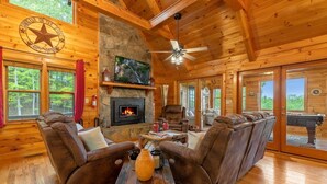 Un intérieur de chalet chaleureux avec une cheminée en pierre, des canapés en cuir, un téléviseur et un ventilateur de plafond.
