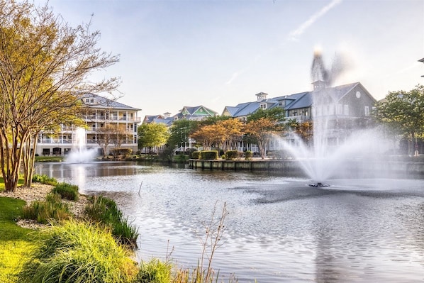 A serene lakeside setting with a fountain, surrounded by multi-story buildings with distinct architectural styles. - Sunset Island 12 Shore Point Drive (Ocean City)