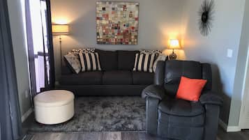 A living room with a dark leather sofa, a black armchair with a red pillow, a beige ottoman, a patterned rug, a floor lamp, and a wall art piece.