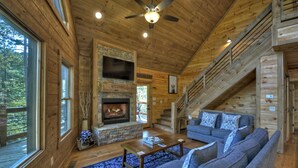 Ein rustikales Holzhaus-Interieur mit Kamin aus Stein, einer Treppe, einer Couch und Blick ins Freie.