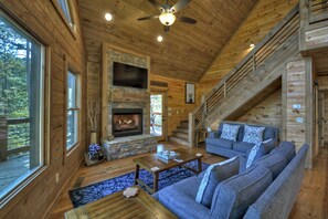 A log cabin interior with a stone fireplace, a staircase, a sofa, and a view of the outdoors.