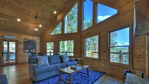 Ein geräumiges Wohnzimmer mit einer großen Couch, einem Couchtisch und einem blauen Teppich mit Muster. Mehrere große Fenster sorgen für natürliches Licht und Ausblicke ins Freie.
