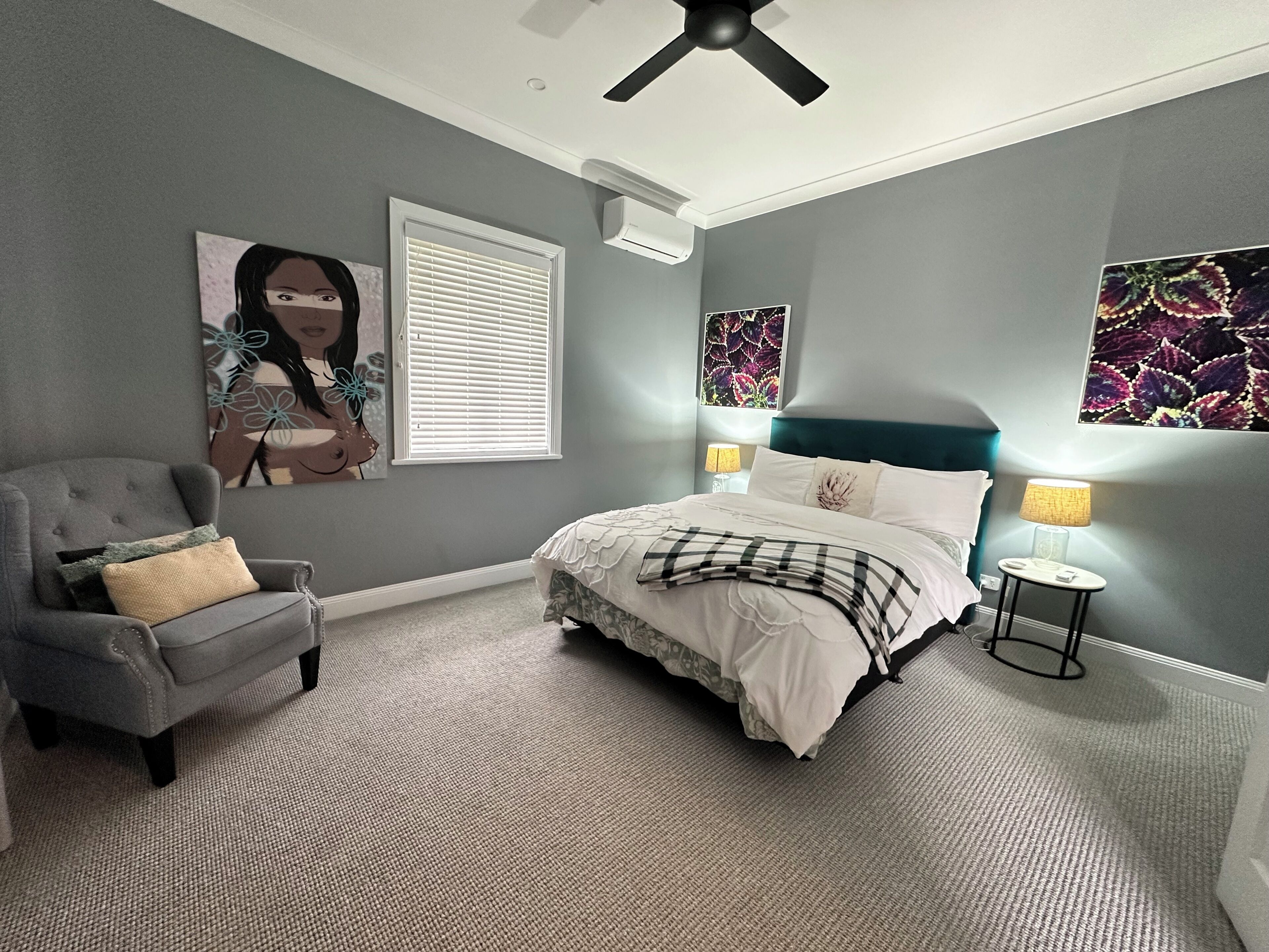 A bedroom with a bed, a chair, a ceiling fan, a window with blinds, and wall art.