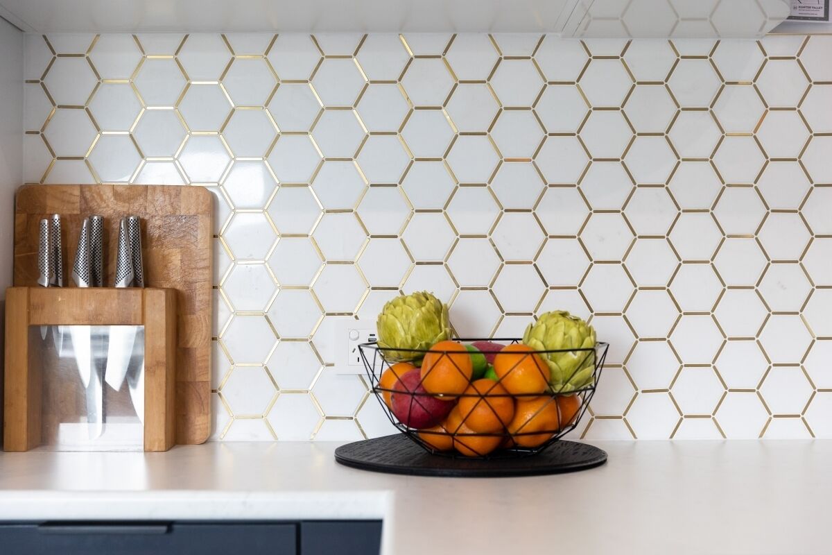 Eine Küchenarbeitsplatte mit einer Obstschale mit Äpfeln, Orangen und Artischocken, ein Holzschneidebrett mit Messern und eine sechseckig gemusterte Spritzschutzwand.