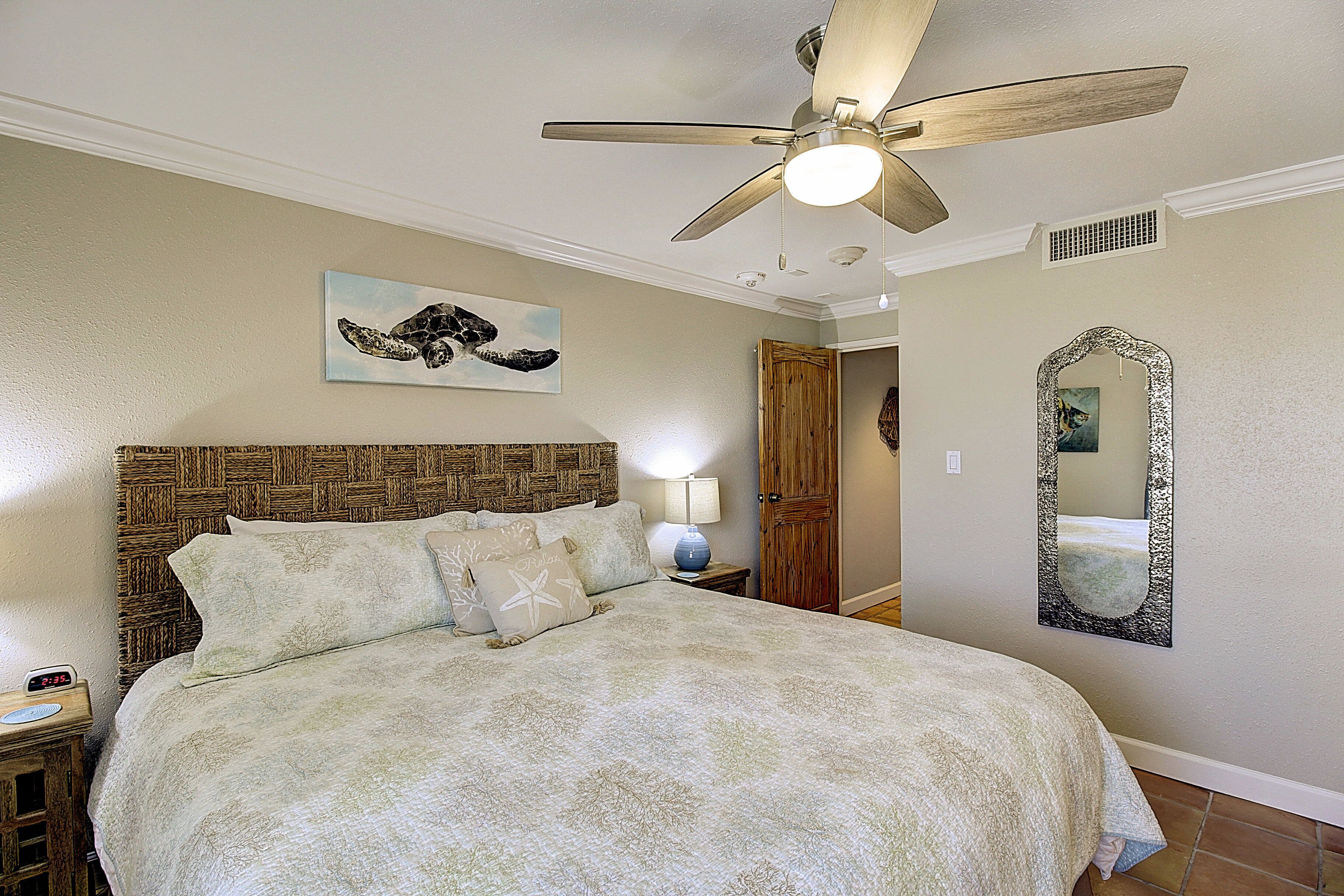 A bedroom with a bed, a ceiling fan, a nightstand, a lamp, a wall-mounted mirror, and a painting.