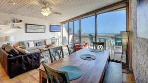 A spacious living area with a wooden dining table, comfortable seating, a ceiling fan, and a large window offering a view of the ocean. - MP218 | Private Entry & Gulf Views | Resort Pools (Port Aransas)