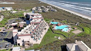 An aerial view of a coastal hotel with multiple buildings, a large swimming pool, and a beach in the background. - MP218 | Private Entry & Gulf Views | Resort Pools (Port Aransas)