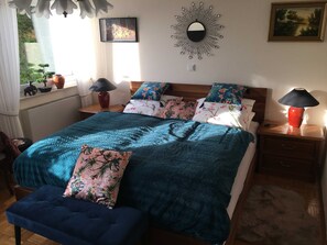 A neatly arranged bedroom with a bed, two nightstands, a wall-mounted mirror, and a window with a view of greenery.