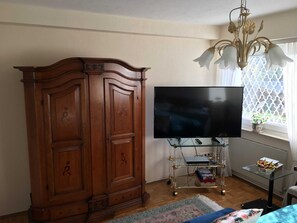 A room with a wooden wardrobe, a flat-screen TV on a glass stand, a chandelier, a window with a patterned curtain, and a small table with items on it.