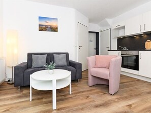 Uma sala de estar moderna com um sofá cinzento, uma poltrona rosa e uma mesa de centro branca. Uma cozinha com armários brancos e bancada preta é visível ao fundo.