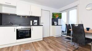 A modern kitchen with white cabinets, a built-in oven, and a coffee machine. There is a dining area with a black chair and a table. - Vacation apartment Abendstern (5) - Holiday apartments in Haus Seekieker in Bensersiel (Esens)