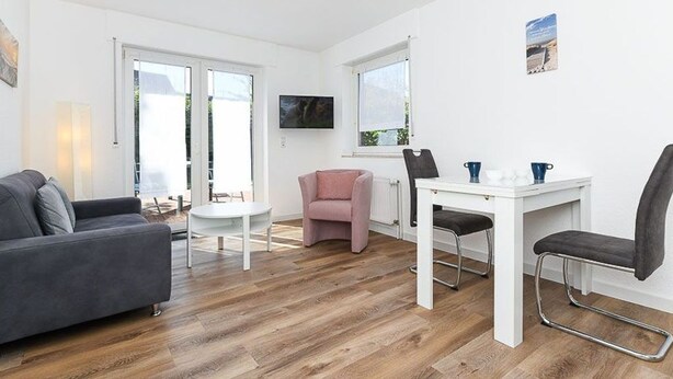 Un salon moderne avec un canapé gris, un fauteuil rose, une table blanche et un téléviseur fixé au mur.
