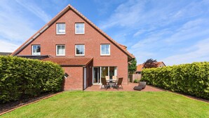 A red brick house with a sloped roof, a patio with a table and chairs, and a well-maintained lawn.