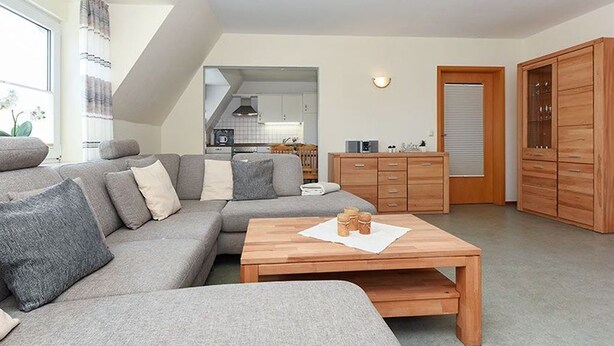 Een moderne woonkamer met een grijze hoekbank, een houten salontafel en een keuken op de achtergrond.