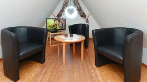 Een kleine, driekantige zolderkamer met twee zwarte leren fauteuils, een houten tafel en een televisie op een standaard.