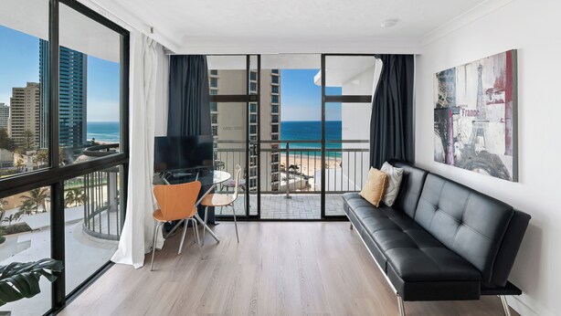 A modern living room with a black leather sofa, a dining table with chairs, and a large window overlooking a beach and ocean.
