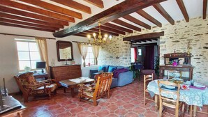 Un interior rústico con vigas de madera a la vista, pared de piedra, sofá azul, mesa de comedor de madera con sillas y una araña.