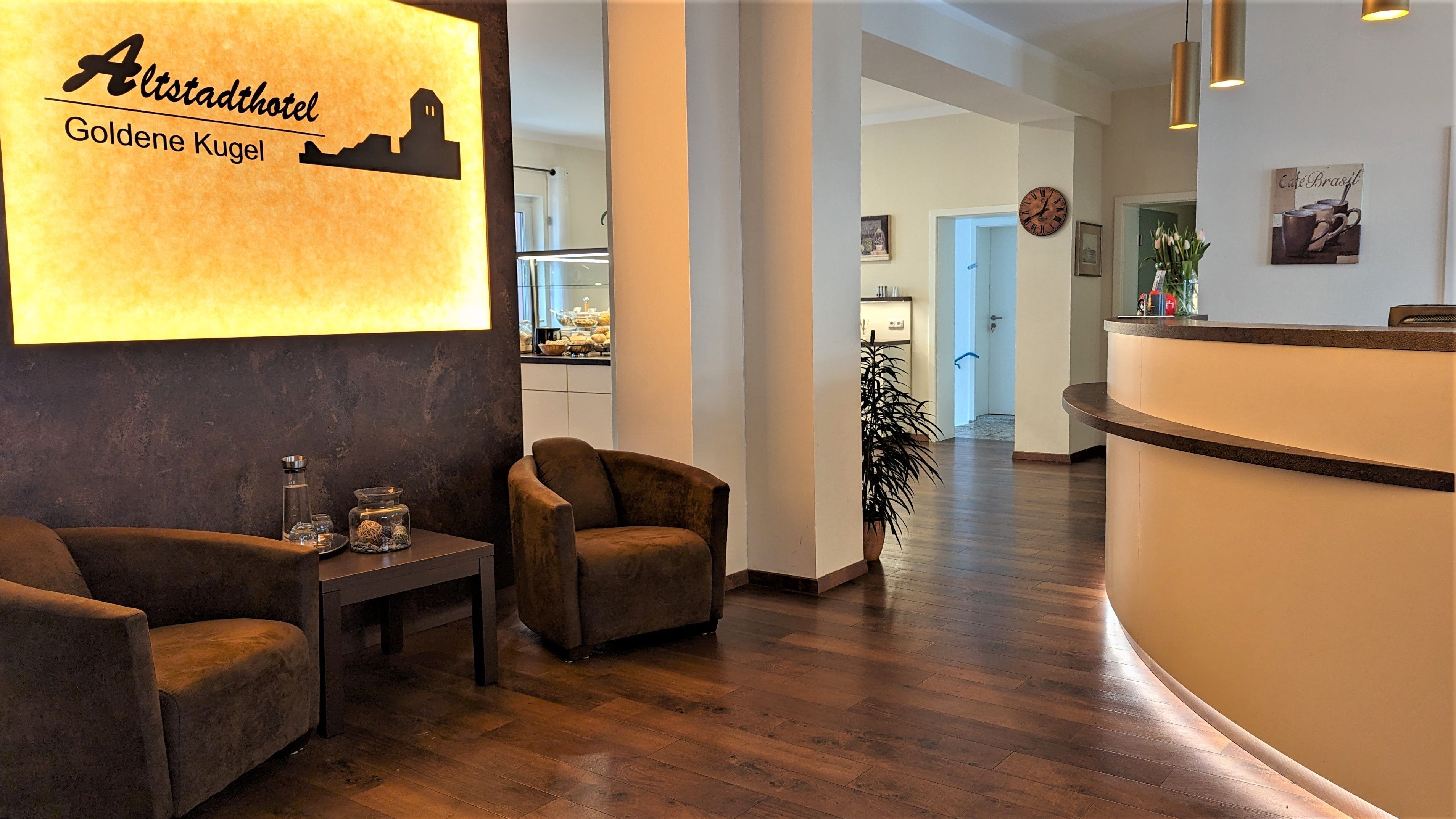 A hotel reception area with a modern design, featuring a reception desk, two armchairs, a small table with decorative items, and a wall-mounted sign with the hotel name and location.
